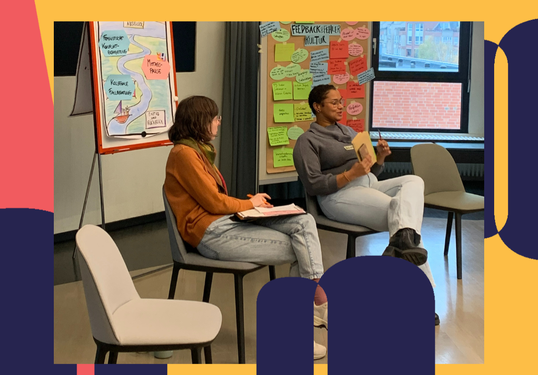 Zwei Teilnehmende sitzen in einem Stuhlkreis und sprechen miteinander während des Workshops der Inklusiven Organisation der Zukunft. Im Hintergrund hängen Pinnwände mit Notizen zu Feedback- und Fehlerkultur sowie ein Flipchart mit Themen wie Kollegiale Fallberatung und feministischer Konfliktbearbeitung. Die Stimmung wirkt konzentriert und offen.