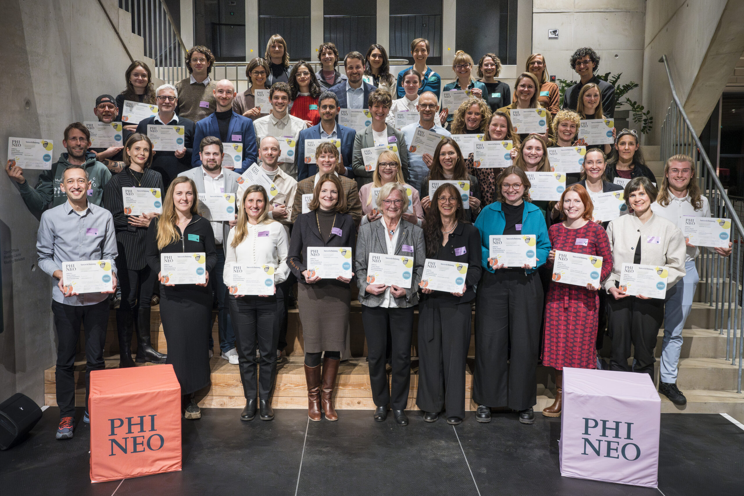 Eine große Gruppe von ausgezeichneten Vertreter*innen zivilgesellschaftlicher Organisationen steht auf mehreren Treppenstufen und hält ihre PHINEO-Wirkt-Siegel-Urkunden gut sichtbar in die Kamera. Die Gruppe ist divers, viele lächeln, einige posieren stolz. Links und rechts im Vordergrund stehen zwei farbige Podeste mit der Aufschrift „PHINEO“. Die Atmosphäre wirkt feierlich und gemeinschaftlich.